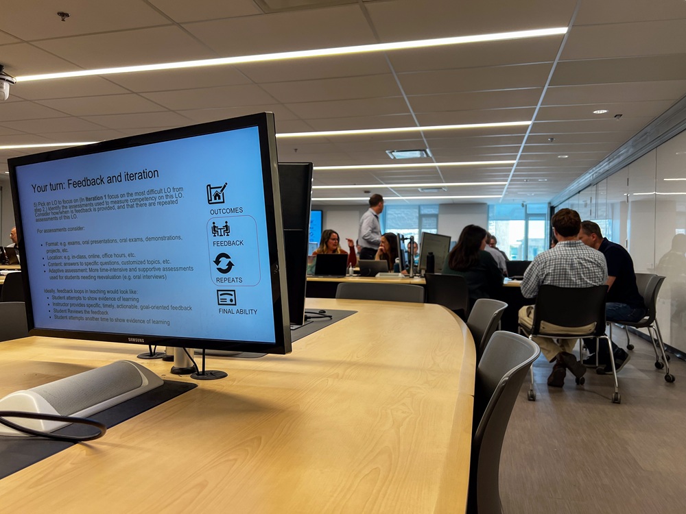 Presentation slide on a monitor in a conference room while people collaborate at tables in the background.