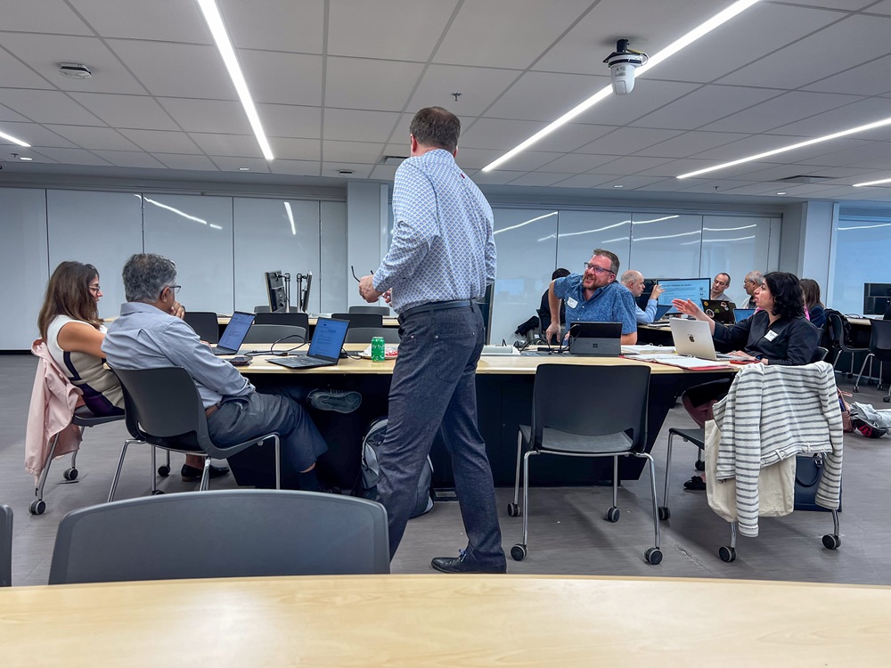 Workshop in a modern conference room, with a facilitator standing as participants work on laptops around a large table.