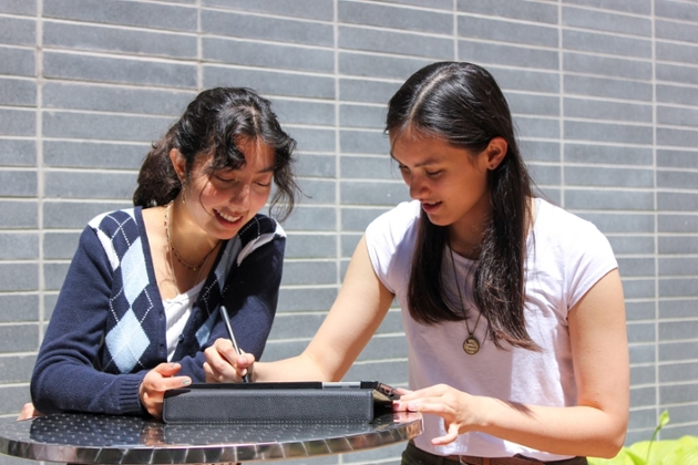 Two students work together at an outdoor table, writing on a tablet with a stylus.
