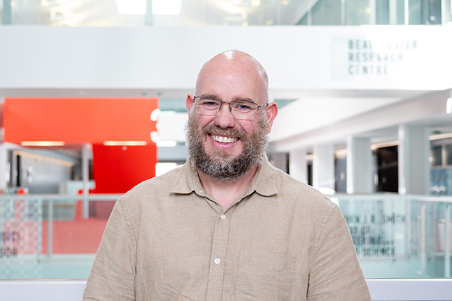 Caucasian man with beard and glasses smiling at camera, it is a corporate headshot. 